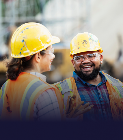 Two individuals in hard hats and vests standing on a construction site, conversing