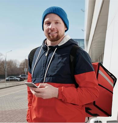 A smiling man in a winter jacket and hat stands outdoors holding his phone with a food delivery bag on his back.