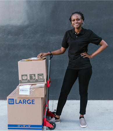 A lady in a black shirt and black pants stands beside a stack of boxes on a trolley