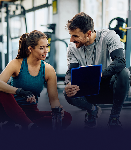 A personal trainer bends down and shows a clipboard to a woman in exercise clothing