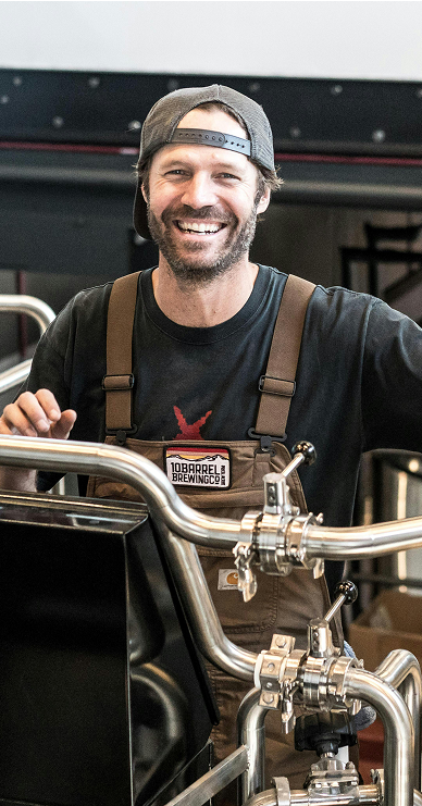 A smiling man in brown overalls standing in front of stainless steel piping and beer brewing tanks