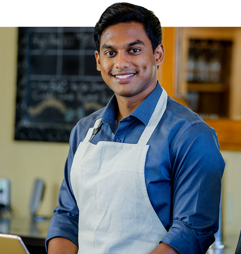 Smiling bearded man wearing a plaid shirt and baseball cap stands in front of a green field with his arms crossed